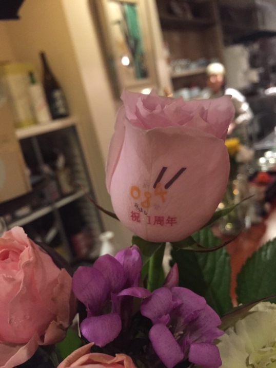 とある飲食店さんへの、オープン1周年お祝いに、お店の常連のお客様から贈られましたお祝いフラワーアレンジメントの1輪のバラに、お店のロゴと『祝 1周年』とのお祝いメッセージをのせております。